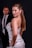 A woman with long, brown hair poses confidently in an elegant, sequined dress. Her expression is alluring, with a slight smile and a direct glance. In the background, a man in a suit observes her from a distance, partially out of focus.