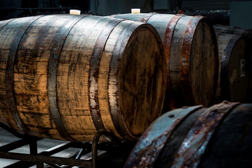 The image depicts several large wooden barrels positioned horizontally on metal racks, in a dimly lit environment. The barrels are weathered and show signs of age with rusted metal hoops.