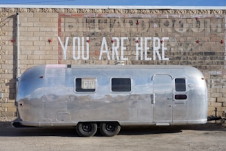An aluminum travel trailer is parked against a brick wall. The wall features a mural with large, bold white letters that read 'YOU ARE HERE.' The trailer's shiny surface reflects its surroundings.