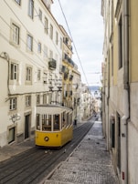 A picturesque street scene features a historic yellow tram ascending a steep, cobblestone street flanked by tall, narrow buildings adorned with balconies. Electrical cables crisscross above, and the street leads to a distant view of the city and sky.