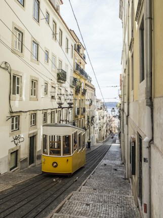 A picturesque street scene features a historic yellow tram ascending a steep, cobblestone street flanked by tall, narrow buildings adorned with balconies. Electrical cables crisscross above, and the street leads to a distant view of the city and sky.