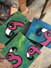 Three wooden panels display painted cartoonish flamingo heads with bright pink colors and large eyes. Each panel has an abstract background, one with a green and another with a blue color. The setting appears to be a workshop or art studio with wood shavings scattered on the floor.