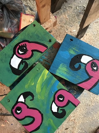 Three wooden panels display painted cartoonish flamingo heads with bright pink colors and large eyes. Each panel has an abstract background, one with a green and another with a blue color. The setting appears to be a workshop or art studio with wood shavings scattered on the floor.