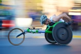 A person is engaged in a wheelchair race, captured in motion. The wheelchair is sleek and aerodynamic, primarily green, with large racing wheels. The racer is wearing a helmet and a sleeveless top, leaning forward with intense concentration. The background is blurred, indicating high speed.