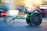 A person is engaged in a wheelchair race, captured in motion. The wheelchair is sleek and aerodynamic, primarily green, with large racing wheels. The racer is wearing a helmet and a sleeveless top, leaning forward with intense concentration. The background is blurred, indicating high speed.
