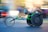 A person is engaged in a wheelchair race, captured in motion. The wheelchair is sleek and aerodynamic, primarily green, with large racing wheels. The racer is wearing a helmet and a sleeveless top, leaning forward with intense concentration. The background is blurred, indicating high speed.