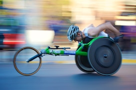 A person is engaged in a wheelchair race, captured in motion. The wheelchair is sleek and aerodynamic, primarily green, with large racing wheels. The racer is wearing a helmet and a sleeveless top, leaning forward with intense concentration. The background is blurred, indicating high speed.