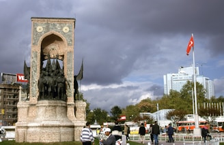 A historic monument with sculptures of figures stands prominently in an urban square. The structure is made of stone with an arched design and is surrounded by people walking. A tall flagpole displays a red flag with a white emblem, situated near modern buildings and lush trees. The sky is overcast with dramatic, dark clouds creating a contrast against the cityscape.