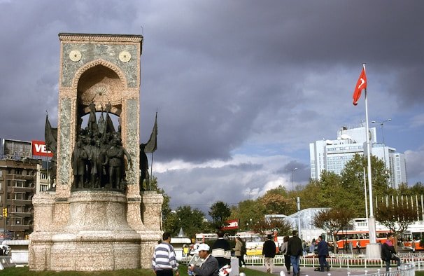 A historic monument with sculptures of figures stands prominently in an urban square. The structure is made of stone with an arched design and is surrounded by people walking. A tall flagpole displays a red flag with a white emblem, situated near modern buildings and lush trees. The sky is overcast with dramatic, dark clouds creating a contrast against the cityscape.