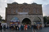 A historic building with an arched entrance and a brick and stone facade stands prominently. The structure features domed roofs and windows with decorative grilles. In front of the building, a crowd of people gathers, some in conversation, suggesting a lively atmosphere. A sign at the entrance reads 'Ünlüoğlu Büfe,' flanked by Pepsi logos.