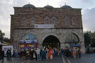 A historic building with an arched entrance and a brick and stone facade stands prominently. The structure features domed roofs and windows with decorative grilles. In front of the building, a crowd of people gathers, some in conversation, suggesting a lively atmosphere. A sign at the entrance reads 'Ünlüoğlu Büfe,' flanked by Pepsi logos.