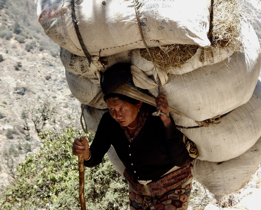 woman carrying sack of hay on back while holding stick