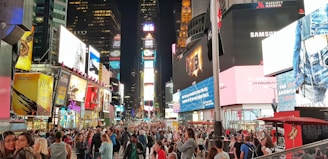 A bustling urban scene filled with a dense crowd of people walking through a brightly lit area lined with large, colorful advertisements on tall buildings. The image features numerous electronic billboards displaying various advertisements, contributing to the vibrant and lively atmosphere.