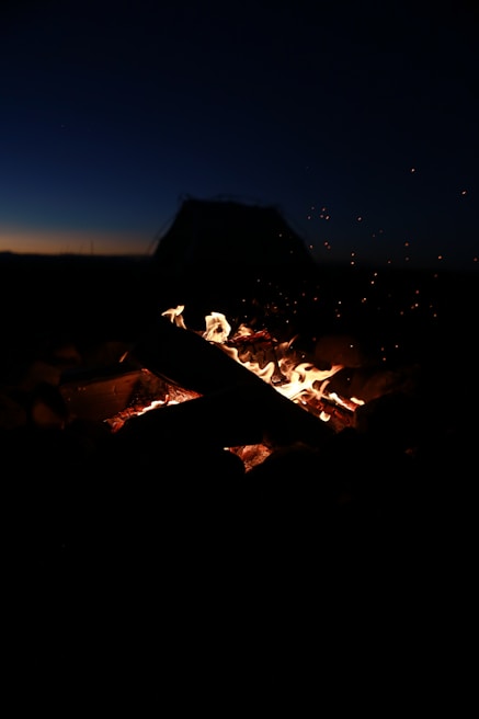 A campfire glowing warmly at dusk with simple camping gear arranged neatly around it on a rocky clearing.