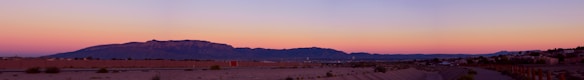 A panoramic view of a desert landscape at sunset with a range of mountains in the background. The sky is a gradient of soft pinks, purples, and blues, creating a serene atmosphere. Sparse vegetation is visible in the foreground, with a few structures or buildings along the horizon.