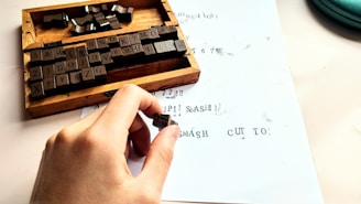 A hand holds a small rubber stamp over a sheet of paper with printed text. Nearby is a wooden box containing various character stamps, including letters, numbers, and symbols. The paper shows a mix of stamped characters, some of which appear slightly smeared or incomplete.