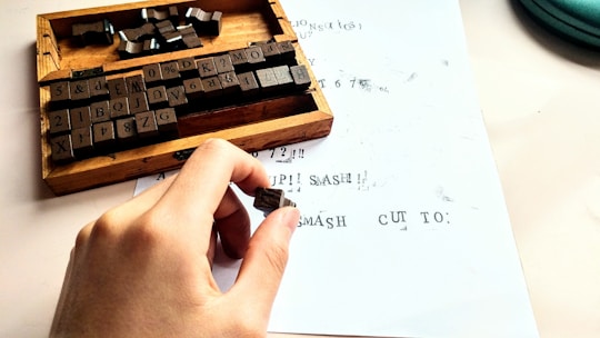 A hand holds a small rubber stamp over a sheet of paper with printed text. Nearby is a wooden box containing various character stamps, including letters, numbers, and symbols. The paper shows a mix of stamped characters, some of which appear slightly smeared or incomplete.