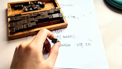 A hand holds a small rubber stamp over a sheet of paper with printed text. Nearby is a wooden box containing various character stamps, including letters, numbers, and symbols. The paper shows a mix of stamped characters, some of which appear slightly smeared or incomplete.