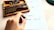 A hand holds a small rubber stamp over a sheet of paper with printed text. Nearby is a wooden box containing various character stamps, including letters, numbers, and symbols. The paper shows a mix of stamped characters, some of which appear slightly smeared or incomplete.