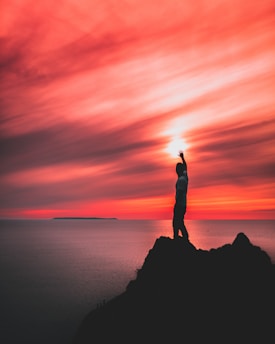 A person stands on the edge of a cliff with an arm raised against a breathtaking sunset sky. The vibrant reds and oranges blend seamlessly with the darker silhouettes of land and sea, creating a dramatic contrast. The ocean stretches out towards the horizon, where an island is faintly visible.
