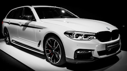A sleek, white, luxury BMW car with sporty design elements, highlighted by black trim and aerodynamic contours. The vehicle is captured in a well-lit setting, emphasizing its glossy finish and intricate headlight design.