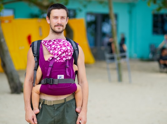 A man is standing outdoors carrying a baby in a baby carrier. The carrier is purple with a patterned fabric covering the baby. The background includes a sandy area and blurred structures, one of which is yellow.