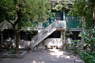 A large, two-story building with green shutters and a central outdoor staircase dominates the scene. The structure is surrounded by lush trees, casting dappled shadows on the paved courtyard below. Several white chairs and tables are arranged under the porch area, suggesting a communal space.