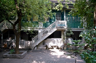 A large, two-story building with green shutters and a central outdoor staircase dominates the scene. The structure is surrounded by lush trees, casting dappled shadows on the paved courtyard below. Several white chairs and tables are arranged under the porch area, suggesting a communal space.
