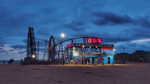 An amusement park scene at dusk featuring a roller coaster illuminated with colorful lights. The sky is a deep blue with scattered clouds, creating a serene backdrop. A small, brightly lit building with signs in a foreign script is situated in front of the coaster. The sandy ground and distant sea add to the coastal atmosphere.