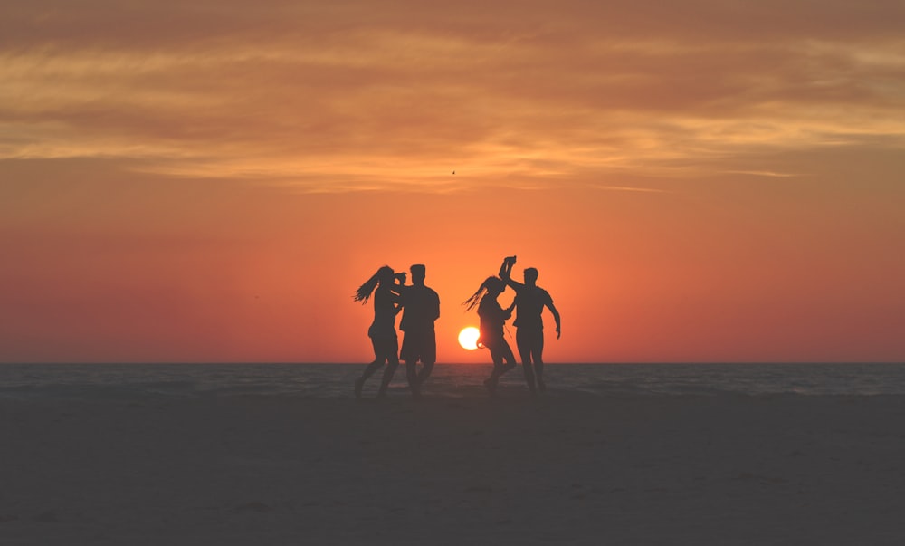 silhouette photo of four people dancing on sands near shoreline