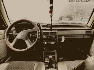 The interior view of a car, featuring a steering wheel with a cover, a dashboard adorned with a hanging charm, and a sticker on the right side depicting a cartoon bird. The windshield shows signs of raindrops and a sticker with text can be seen at the top right corner.