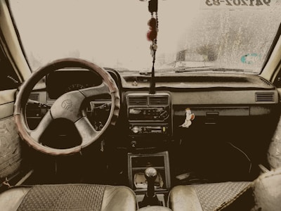 The interior view of a car, featuring a steering wheel with a cover, a dashboard adorned with a hanging charm, and a sticker on the right side depicting a cartoon bird. The windshield shows signs of raindrops and a sticker with text can be seen at the top right corner.
