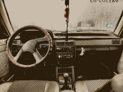 The interior view of a car, featuring a steering wheel with a cover, a dashboard adorned with a hanging charm, and a sticker on the right side depicting a cartoon bird. The windshield shows signs of raindrops and a sticker with text can be seen at the top right corner.