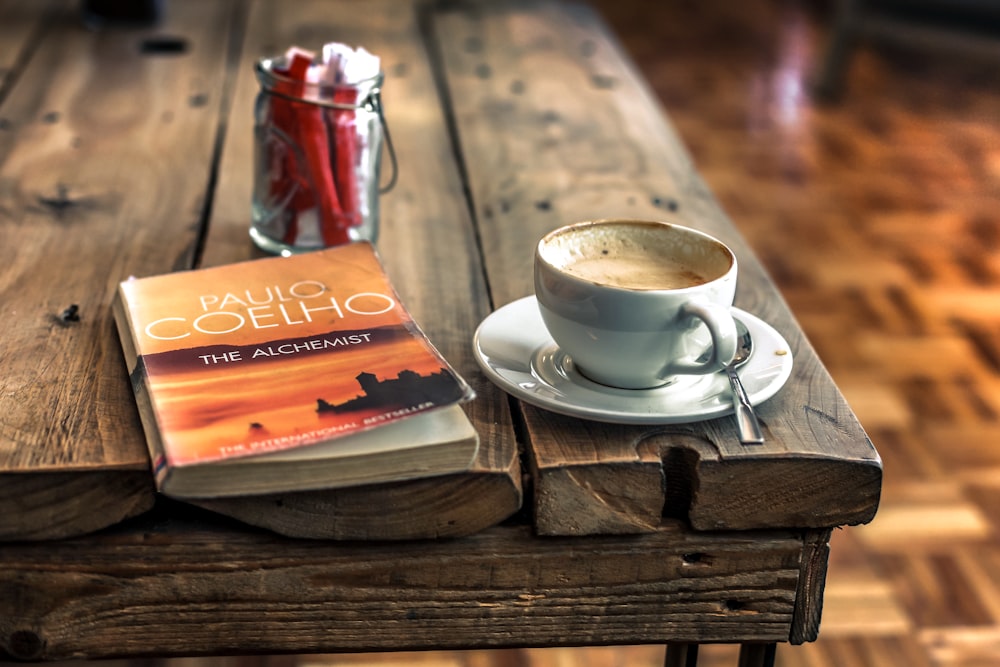 white and gray ceramic mug filled with coffee on white ceramic saucer with gray stainless steel spoon on brown wooden table