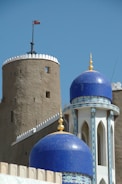 A historic fort tower with a flag flies beside a mosque featuring blue domes adorned with intricate mosaic patterns and golden finials. The architecture combines medieval military and Islamic elements, set against a clear blue sky.