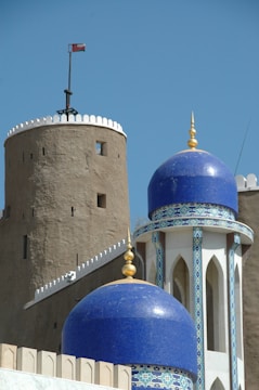 A historic fort tower with a flag flies beside a mosque featuring blue domes adorned with intricate mosaic patterns and golden finials. The architecture combines medieval military and Islamic elements, set against a clear blue sky.