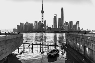 A monochromatic cityscape features the iconic skyline of skyscrapers reflecting on a river with a small boat in the foreground. The skyline is captured between two stone walls, creating a framed perspective.