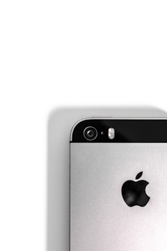 A close-up view of the back of a silver smartphone, featuring a camera lens, flash, and the iconic apple logo. The upper portion of the phone contains a black strip that houses the camera and flash components, while the rest of the body is metallic.