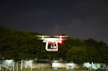 A white drone is flying in the air with its red and green lights illuminated, set against a backdrop of dark trees and a cloudy night sky. The drone's rotors are in motion, indicating active flight.
