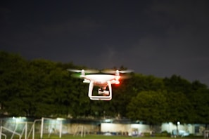 A white drone is flying in the air with its red and green lights illuminated, set against a backdrop of dark trees and a cloudy night sky. The drone's rotors are in motion, indicating active flight.