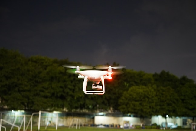 A white drone is flying in the air with its red and green lights illuminated, set against a backdrop of dark trees and a cloudy night sky. The drone's rotors are in motion, indicating active flight.