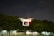 A white drone is flying in the air with its red and green lights illuminated, set against a backdrop of dark trees and a cloudy night sky. The drone's rotors are in motion, indicating active flight.