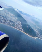 An aerial view of a coastline with a sprawling cityscape adjacent to lush green hills. The ocean is visible below, with the airplane's engine and part of the wing in the foreground. Clouds are scattered above, adding depth to the scene.