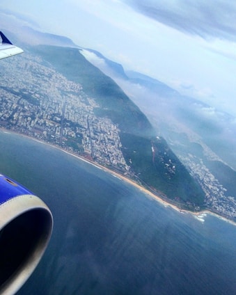An aerial view of a coastline with a sprawling cityscape adjacent to lush green hills. The ocean is visible below, with the airplane's engine and part of the wing in the foreground. Clouds are scattered above, adding depth to the scene.