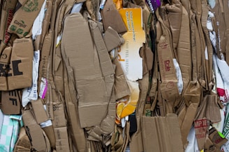 A stack of flattened and tightly packed cardboard boxes and packaging materials, displaying various printed labels, logos, and text in multiple colors. The cardboard is crumpled and layered, showing visible wear and tear, and some pieces have torn edges.