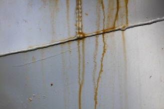 Rusty stains run down a pale gray metallic surface with visible seams and welding marks, indicating signs of corrosion.
