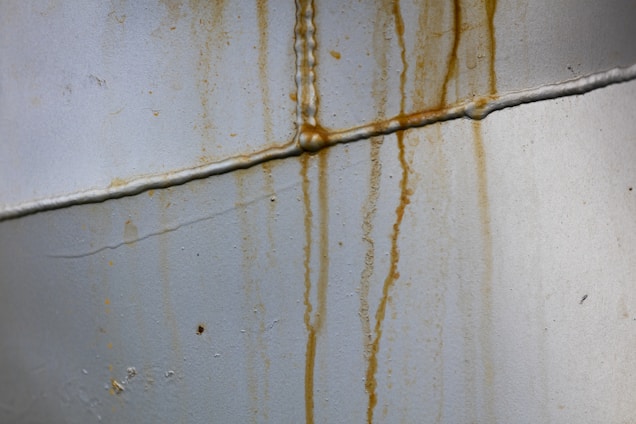 Rusty stains run down a pale gray metallic surface with visible seams and welding marks, indicating signs of corrosion.