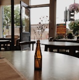 A small brown glass bottle with delicate white flowers sits on a table in a cafe. The background shows an open door leading outside, with soft natural light filtering through. There are chairs and tables scattered around, and a colorful poster is visible near the entrance, adding vibrancy to the otherwise muted tones.