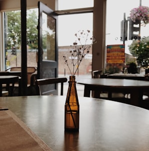 A small brown glass bottle with delicate white flowers sits on a table in a cafe. The background shows an open door leading outside, with soft natural light filtering through. There are chairs and tables scattered around, and a colorful poster is visible near the entrance, adding vibrancy to the otherwise muted tones.