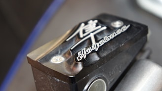 A close-up of a motorcycle part with embossed text and symbols. The text 'Hayabusa' is prominently featured alongside a stylized Japanese character. The part has a metallic, angular design with a black finish and highlights visible on its surface.